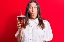 Beautiful Brunette Woman Drinking Cola Refreshment Beverage Using Straw Over Red Background Scared And Amazed With Open Mouth For Surprise, Disbelief Face