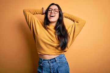 Young brunette woman wearing glasses and casual sweater over yellow isolated background relaxing and stretching, arms and hands behind head and neck smiling happy