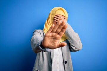 Wall Mural - Middle age brunette business woman wearing muslim traditional hijab over blue background covering eyes with hands and doing stop gesture with sad and fear expression. Embarrassed and negative concept.