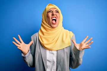 Wall Mural - Middle age brunette business woman wearing muslim traditional hijab over blue background crazy and mad shouting and yelling with aggressive expression and arms raised. Frustration concept.