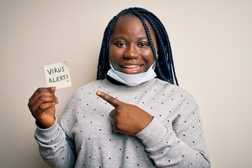 Sticker - African american woman wearing medical mask holding reminder with virus alert message very happy pointing with hand and finger