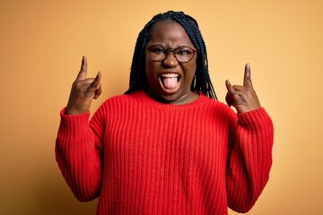 Sticker - African american plus size woman with braids wearing casual sweater over yellow background shouting with crazy expression doing rock symbol with hands up. Music star. Heavy concept.