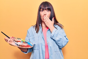 Young plus size woman holding paintbrush and palette covering mouth with hand, shocked and afraid for mistake. surprised expression