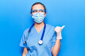 Wall Mural - Young blonde woman wearing doctor uniform and coronavirus protection holding help reminder pointing thumb up to the side smiling happy with open mouth