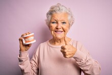 Senior Beautiful Grey-haired Woman Holding Plastic Denture Teeth Over Pink Background Happy With Big Smile Doing Ok Sign, Thumb Up With Fingers, Excellent Sign