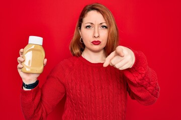 Poster - Beautiful redhead woman holding bottle of mustard sauce condiment over red background pointing with finger to the camera and to you, hand sign, positive and confident gesture from the front