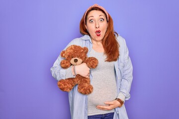 Poster - Young redhead pregnant woman expecting baby holding teddy bear over purple background scared in shock with a surprise face, afraid and excited with fear expression