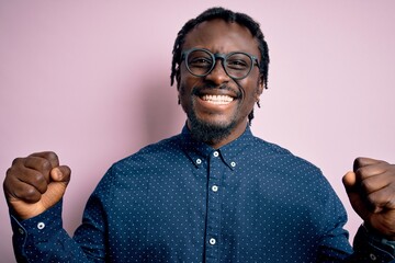 Poster - Young handsome african american man wearing casual shirt and glasses over pink background celebrating surprised and amazed for success with arms raised and open eyes. Winner concept.