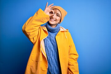 Poster - Young blonde woman with short hair wearing rain coat for rainy weather over blue background doing ok gesture with hand smiling, eye looking through fingers with happy face.