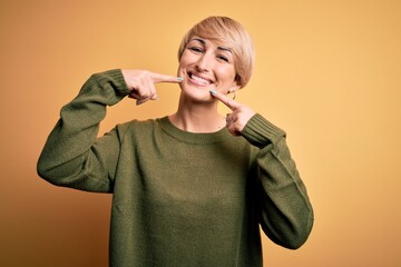 Poster - Young blonde woman with modern short hair wearing casual sweater over yellow background smiling cheerful showing and pointing with fingers teeth and mouth. Dental health concept.