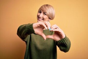 Wall Mural - Young blonde woman with modern short hair wearing casual sweater over yellow background smiling in love doing heart symbol shape with hands. Romantic concept.