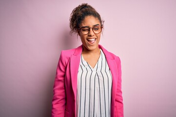 Wall Mural - Beautiful african american businesswoman wearing jacket and glasses over pink background winking looking at the camera with sexy expression, cheerful and happy face.