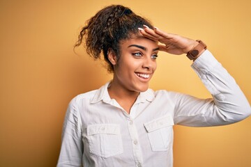 Sticker - Young beautiful african american girl wearing casual shirt standing over yellow background very happy and smiling looking far away with hand over head. Searching concept.