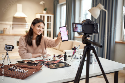 Spread your charm. Beauty blogger woman filming, advertising app on camera, holding smartphone. Makeup influencer asian girl recording cosmetics product review at home