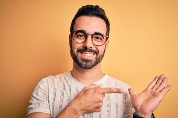 Canvas Print - Young handsome man with beard wearing casual t-shirt and glasses over yellow background amazed and smiling to the camera while presenting with hand and pointing with finger.