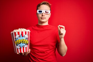 Wall Mural - Young handsome redhead man watching 3d glasses eating popcorn snack over red background with a confident expression on smart face thinking serious