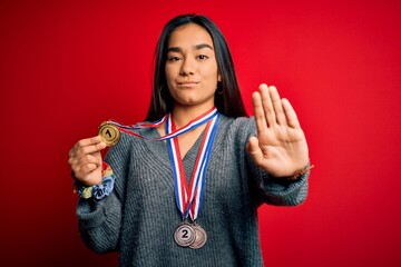 Wall Mural - Young beautiful successful asian woman wearing medals standing over red background with open hand doing stop sign with serious and confident expression, defense gesture