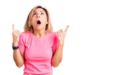 Young blonde woman wearing sportswear amazed and surprised looking up and pointing with fingers and raised arms.