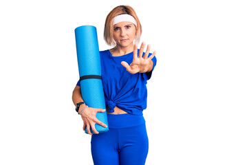 Sticker - Young blonde woman holding yoga mat with open hand doing stop sign with serious and confident expression, defense gesture