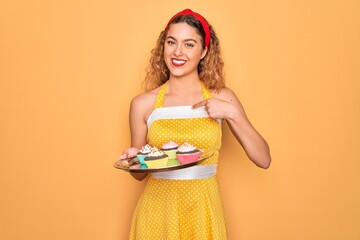 Wall Mural - Beautiful pin-up woman with blue eyes holding chocolate cupcakes over yellow background with surprise face pointing finger to himself