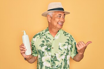 Poster - Middle age senior grey-haired man wearing summer hat holding sun protection cream pointing and showing with thumb up to the side with happy face smiling