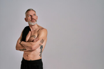 Wall Mural - Middle aged muscular man took off black t shirt, showing his body, looking aside while posing in studio over grey background