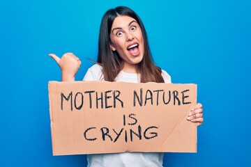 Young woman asking for environment care holding banner with mother nature is crying message pointing thumb up to the side smiling happy with open mouth