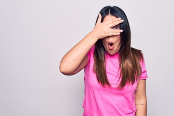 Poster - Young beautiful brunette woman wearing casual pink t-shirt standing over white background peeking in shock covering face and eyes with hand, looking through fingers with embarrassed expression.