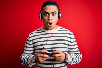 Young brazilian gamer man playing video game using joystick and headphones scared in shock with a surprise face, afraid and excited with fear expression