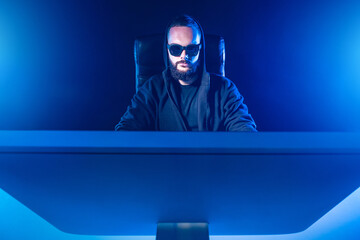 Poster - The concept of blogging. A man in black glasses and a hoodie behind a monitor. Blogger on a blue background. Place for text. Working at the computer. Creative photo with a blogger.