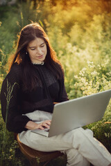 Wall Mural - New office concept. Fashionable elegant girl working on laptop and sitting on rustic chair in warm sunset light in summer field. Remote work with social distancing and safety protocols