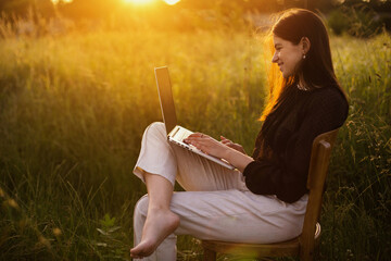 Wall Mural - New office concept. Fashionable elegant girl working on laptop and sitting on rustic chair  in sunny summer field at sunset.  Remote work with social distancing and safety protocols