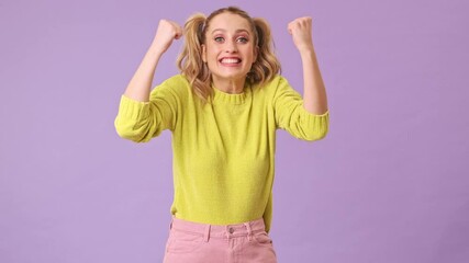 Sticker - Happy amazed young blonde girl celebrates victory raising arms in an isolated studio on a purple background