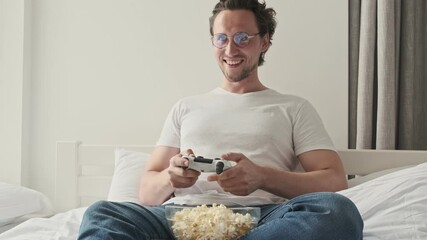 Wall Mural - Happy handsome man in eyeglasses playing on console with gamepad while sitting with popcorn on sofa at home