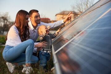 Wall Mural - Side view shot of a young modern family with a little son getting acquainted with solar panel on a sunny day, green alternative energy concept