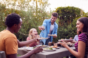 Multi-Cultural Friends At Home Sitting At Table Enjoying Food At Summer Garden Party