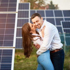 Wall Mural - Close-up shot of a young couple standing between solar panels, woman is kissing a man in a cheek, bent backwards, man hugging her, bent forward