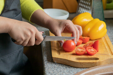 Wall Mural - Close up view of female cook slices cherry tomatoes on wooden board colorful vegetables on background, woman cooking fresh low-calorie salad in kitchen. Healthy food, diet, organic farm products