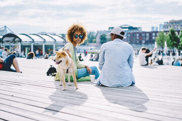 Stylish african american hipster girl in cool sunglasses stroke dog while sitting in urban setting and spending free time together with boyfriend.Dark skinned young man and woman resting i modern city