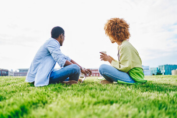 Back view of two african american hipster friends dressed in casual wear sitting on green grass in park.Couple in love enjoying coffee beverage and resting in urban setting in sunny day