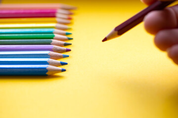 Colored pencils in a row in rainbow colors, hand with blue colored pencil draws on orange paper, retro background