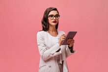 Serious Business Lady With Bright Red Lips In Beige Stylish Outfit And Eyeglasses Holds Tablet On Pink Isolated Background..