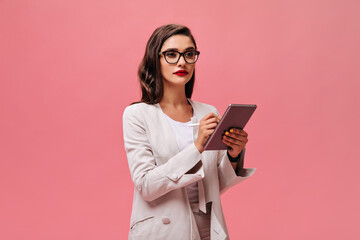 Serious business lady with bright red lips in beige stylish outfit and eyeglasses holds tablet on pink isolated background..