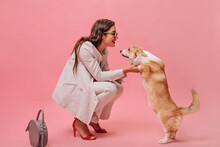 Woman In Beige Suit Plays With Dog On Pink Background. Cute Beautiful Girl With Glasses And Red Heels Looks At Corgi And Smiles..