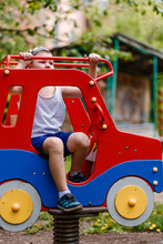 A Boy In Shorts And A T-shirt Sits On A Children's Red Car In The Park In The Summer.