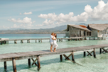 Wall Mural - Couple on the beach at tropical resort. Travel honeymoon concept. Happy loving couple in white clothes hug each other while stand on wooden jetty