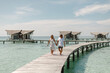 © Yevhenii - Happy loving couple walking on summer tropical beach on wooden jetty with overwater wooden bungalow on background