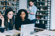 © BullRun - Serious multiracial women looking at laptop while smart man reading book at bookshelves
