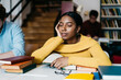 © BullRun - Tired african american student dressed in casual wear sleeping while sitting at desktop with many literature books for exam preparation in modern classroom during conducting boring studying lesson