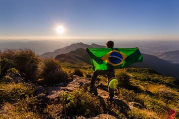 Canvas Print - Young male holding a flag of Brazil and standing on top of a hill